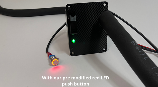Black carbon fiber textured motorsport hydration module showing green indicator LED with a RED LED push button next to it connected to a wire to the drink module. Insulated black tubing routes in and out of the unit on a white background with a text overlay saying "With our pre modified red LED push button"