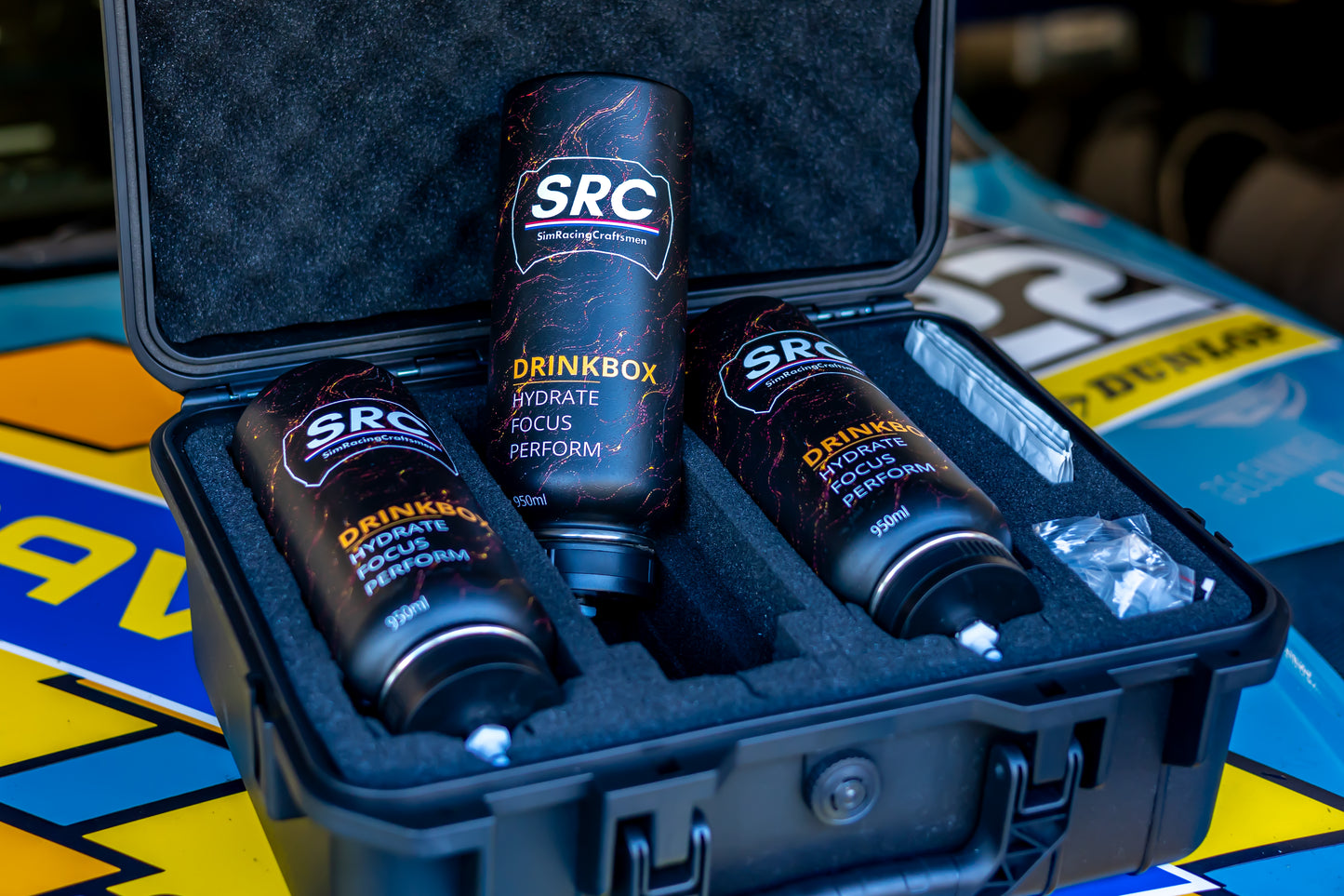 Three black SRC Drinkbox motorsport quick change drink bottles arranged inside an open black hard case with foam lining, photographed close-up on a branded blue and yellow surface.