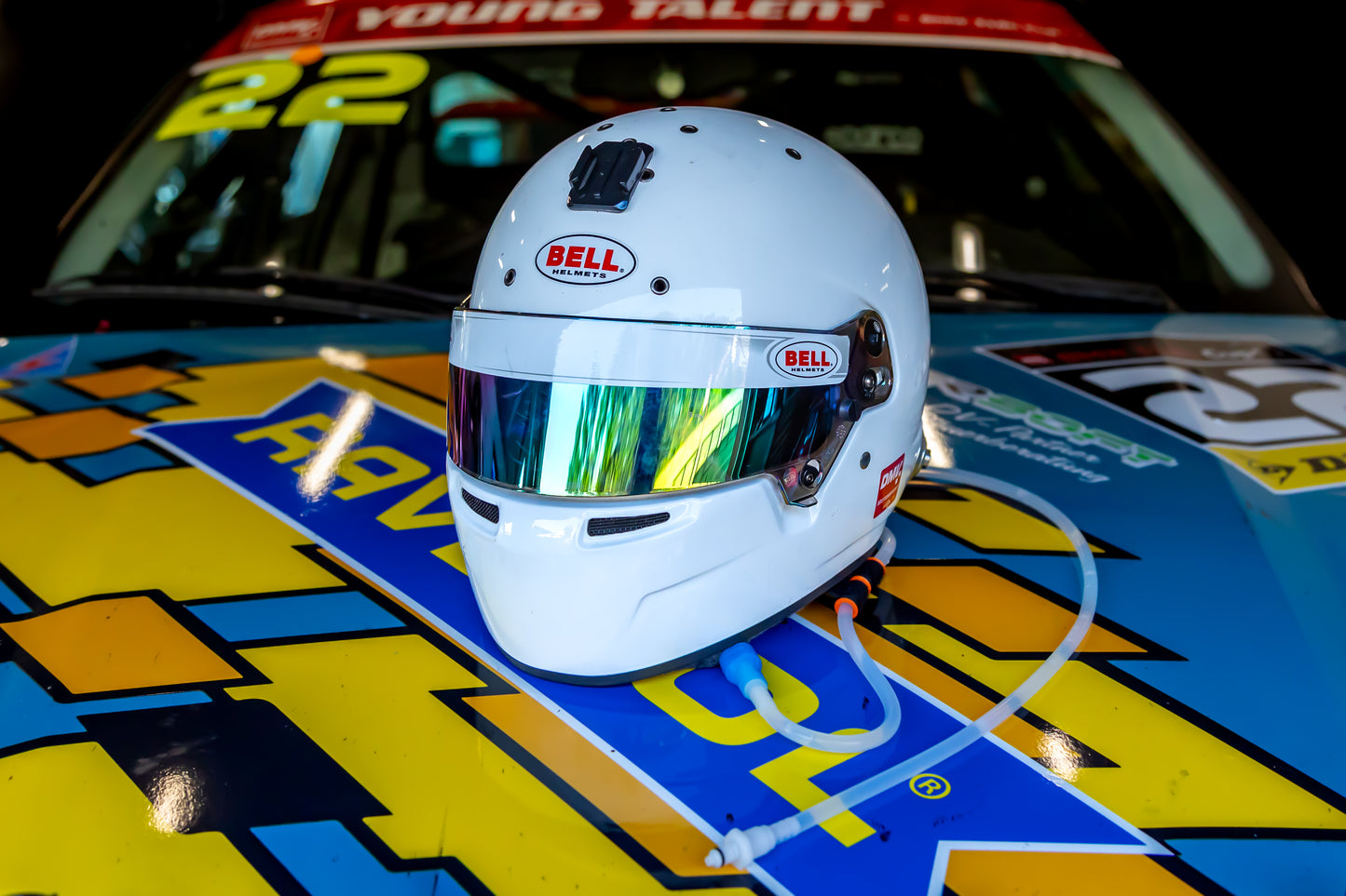 A white Bell racing helmet with a reflective visor resting on the hood of a brightly colored race car at night. with motorsport hydration tubing hanging from the helmet