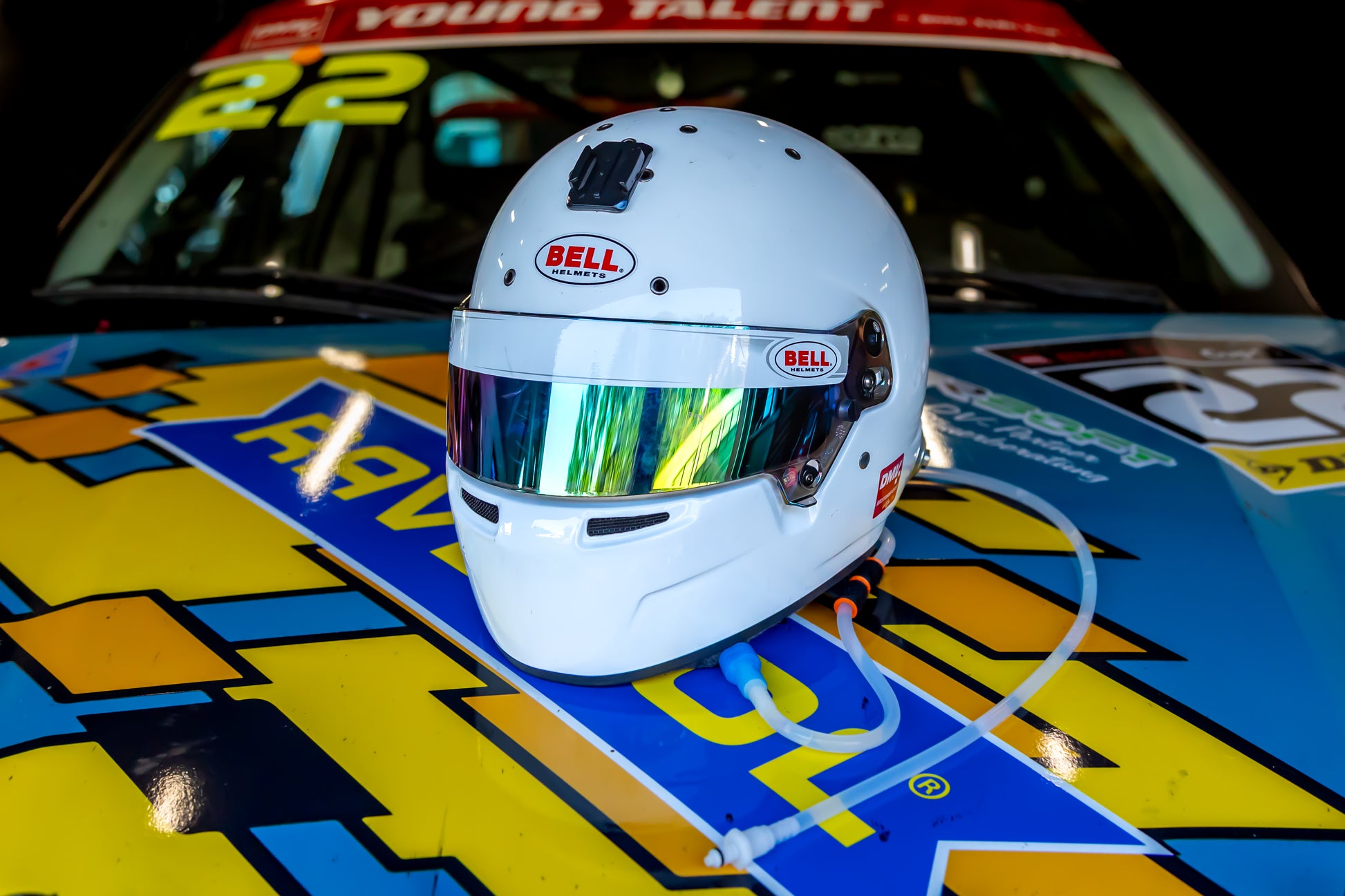 A white Bell racing helmet with a reflective visor resting on the hood of a brightly colored race car at night. with motorsport hydration tubing hanging from the helmet