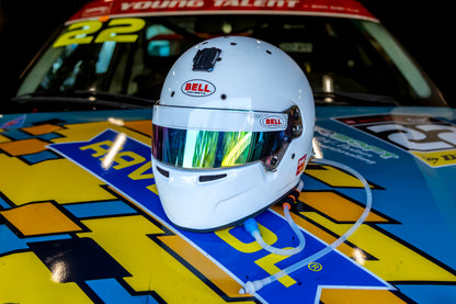 A white Bell racing helmet with a reflective visor resting on the hood of a brightly colored race car at night. with motorsport hydration tubing hanging from the helmet