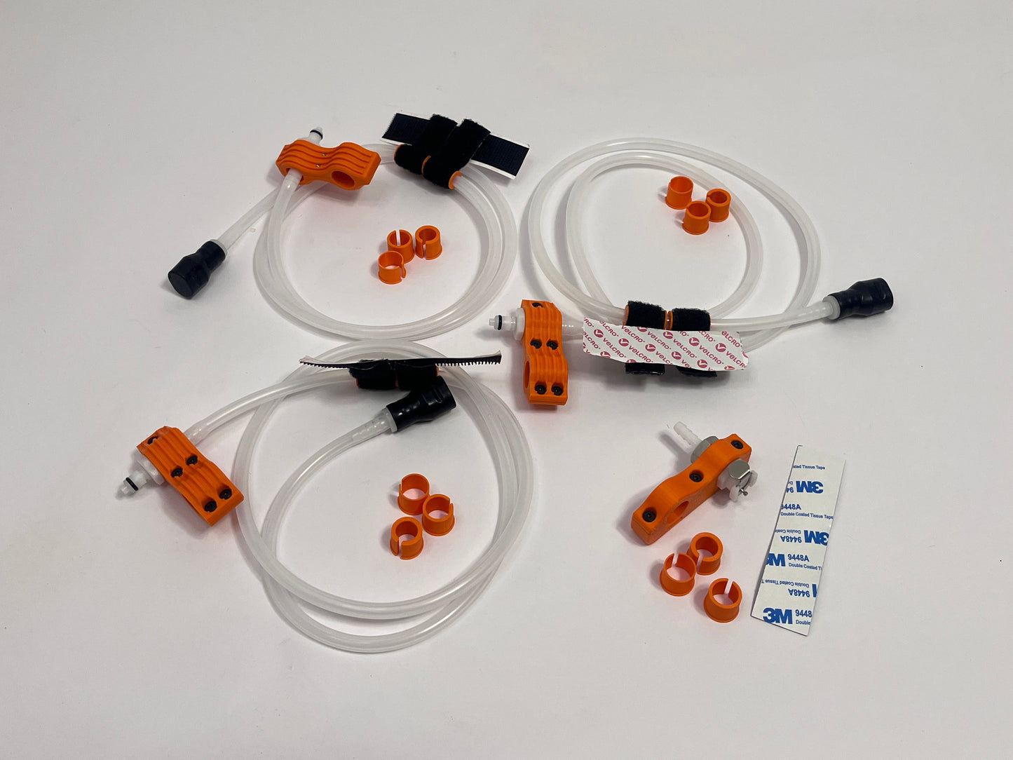 three driver side motorsport helmet drinking kits showing per set orange dry break fitting holder, 1m silicone tubing connected to it with 3 spacers shown loosely. black silicone bite valve is connected to the tube and velcro positioners connected to the tubing. also a car side connector in orange shown with dry break fitting installed, 3m backed rubber and 3 orange radio plug spacers on a light gray background