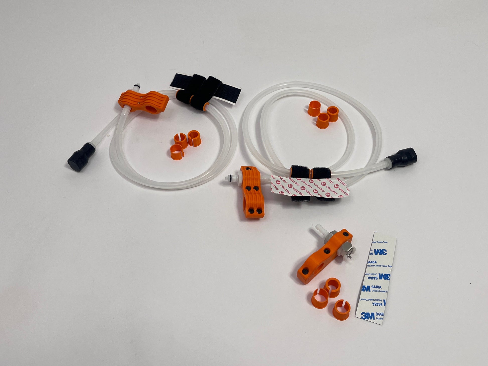 two driver side racing hydration kits showing per set orange dry break fitting holder, 1m silicone tubing connected to it with 3 spacers shown loosely. black silicone bite valve is connected to the tube and velcro positioners connected to the tubing. also a car side connector in orange shown with dry break fitting installed, 3m backed rubber and 3 orange radio plug spacers on a light gray background