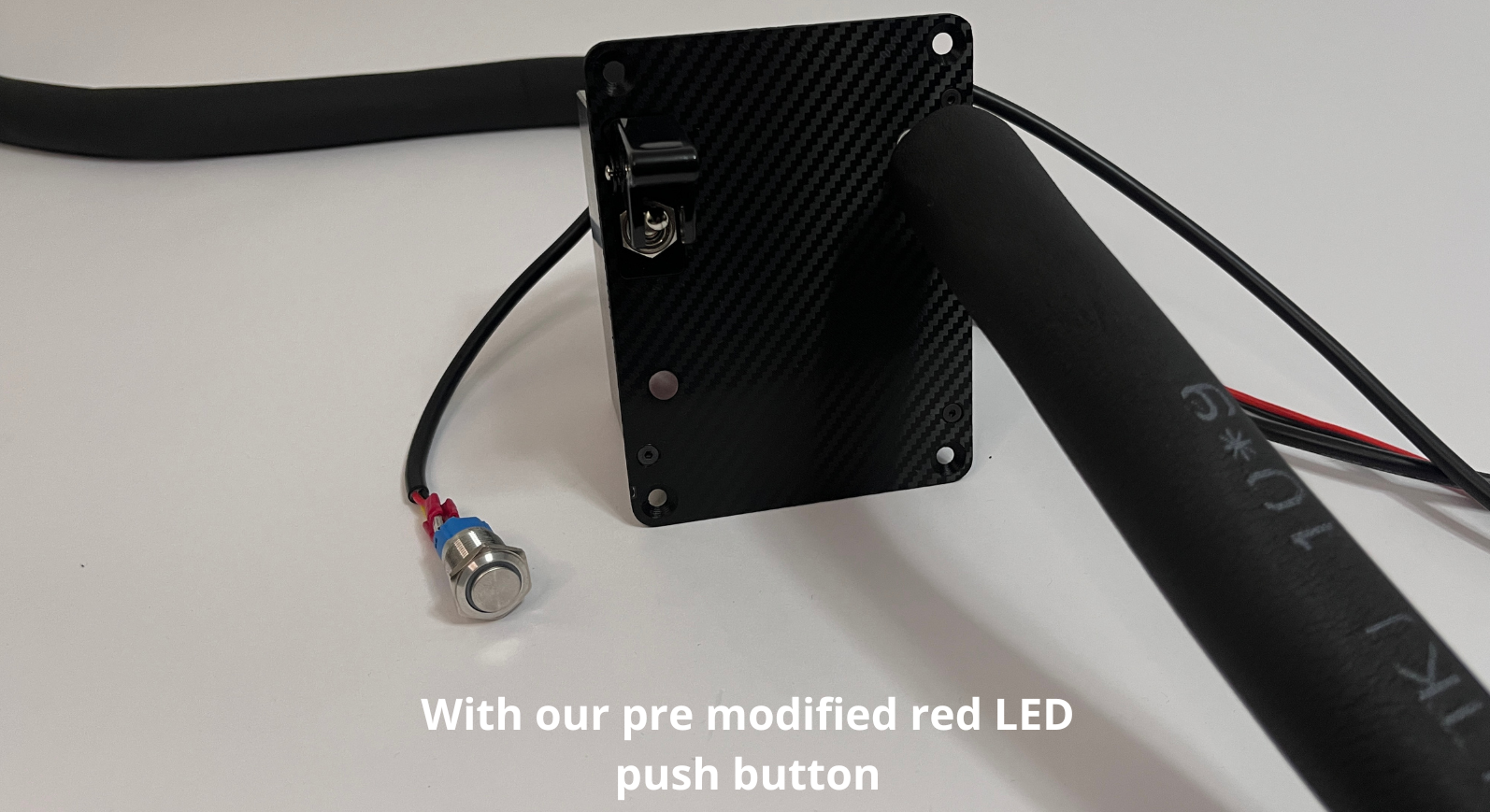 A Black carbon fiber textured racing water hydration pump box showing green indicator LED with a off push button next to it connected to a wire to the drink module. Insulated black tubing routes in and out of the unit on a white background with a text overlay saying "With our pre modified red LED push button"