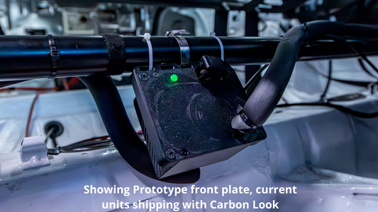 A black automotive racing hydration drink module with a green indicator light is mounted on a roll bar inside a racing car cabin. White text at the bottom reads 'Showing Prototype front plate, current units shipping with Carbon Look'.